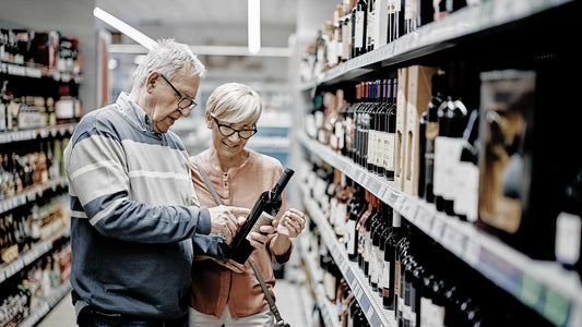 Ein altes Ehepaar schaut sich eine Flasche Wein im Supermarkt an, umgeben von Regalen.