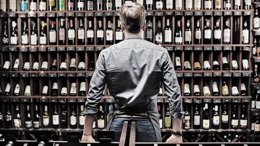 Ein Mann steht vor einem Weinregal in einer Weinhandlung mit viel Flaschen Bordeaux-Wein und verschiedenen Jahrgängen