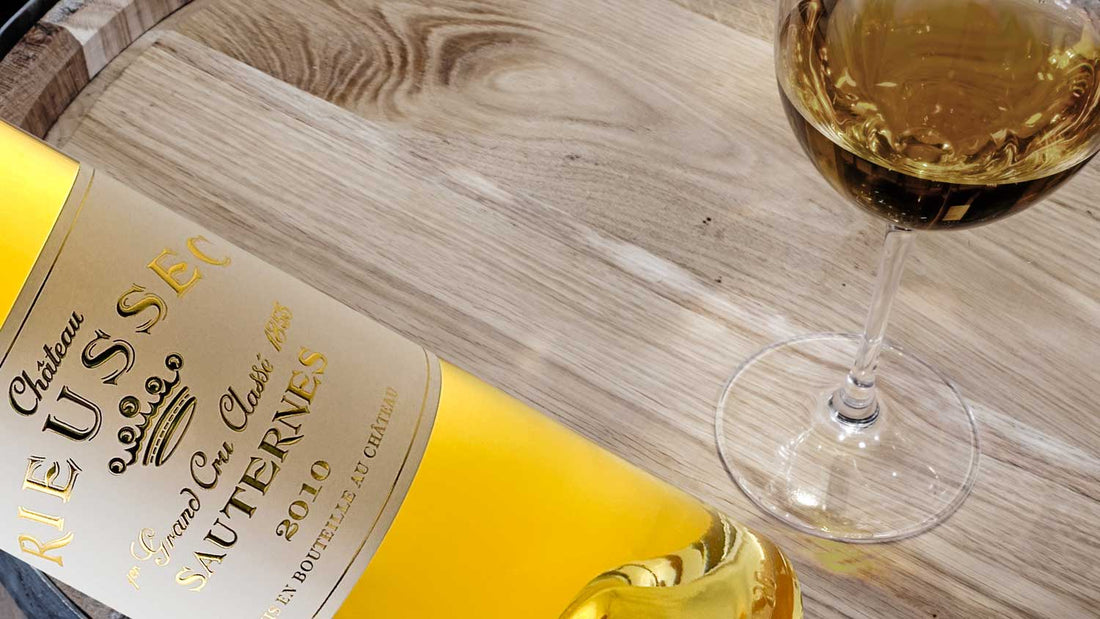 Eine Flasche Dessertwein von Chateau Rieussec im Vordergrund, daneben ein Glas Wein auf einem Holzfass
