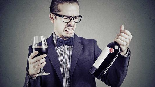 Ein Mann hält mit angewidertem Gesichtsausdruck eine Flasche Wein und ein volles Weinglas in der Hand.