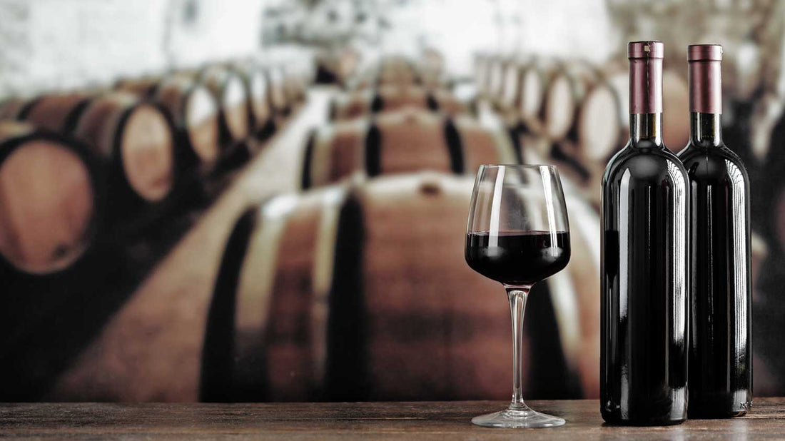 Zwei Weinflaschen und ein Weinglas mit Rotwein stehen vor Weinfässern in einem Weinkeller