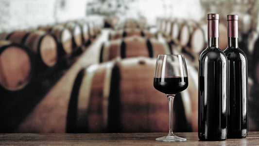 Zwei Weinflaschen und ein Weinglas mit Rotwein stehen vor Weinfässern in einem Weinkeller