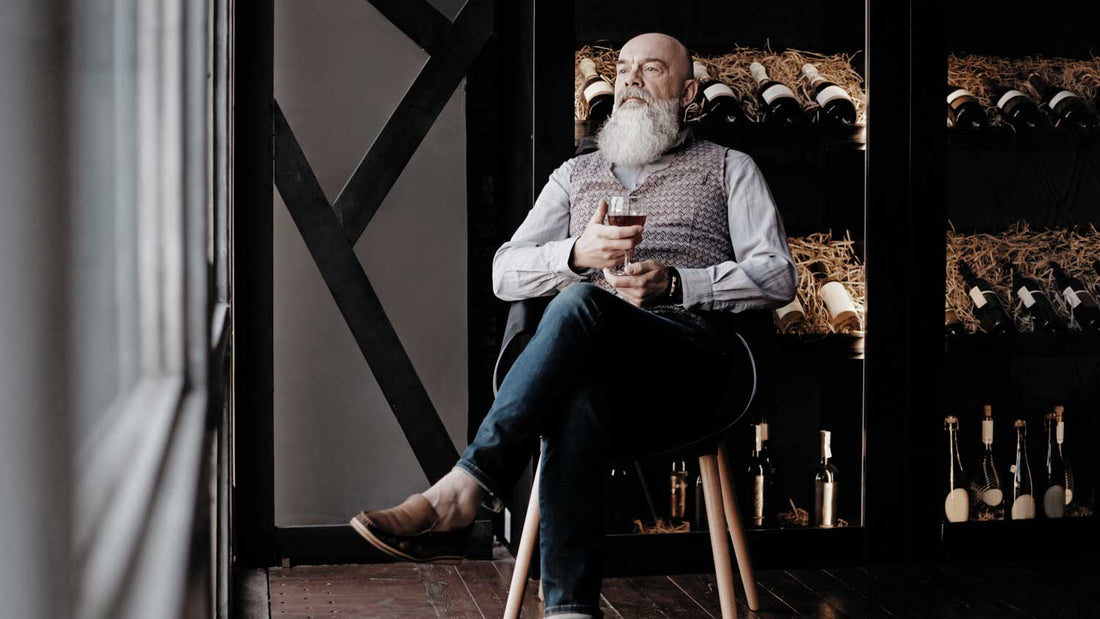 Ein Weinsammler sitzt auf einem Stuhl vor einem Weinregal und sieht nachdenklich aus.