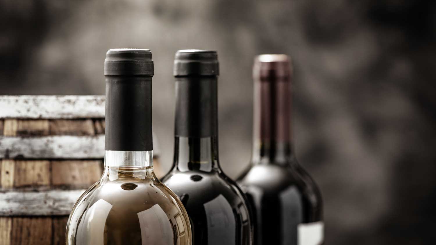 Das Bild zeigt eine Flasche Weißwein und zwei Flaschen Rotwein, die vor einem Weinfass stehen.