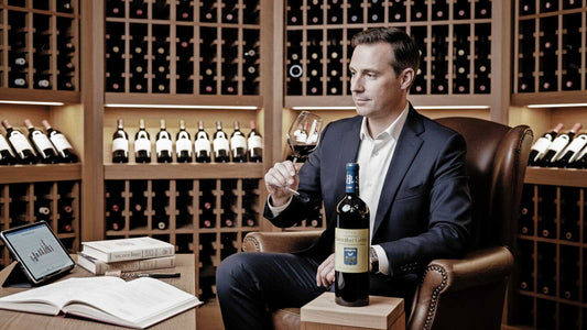 Mann im Anzug sitzt in Ledersessel in Weinkeller, hält Weinglas, vor ihm steht große Weinflasche, im Hintergrund Weinregale mit Flaschen.