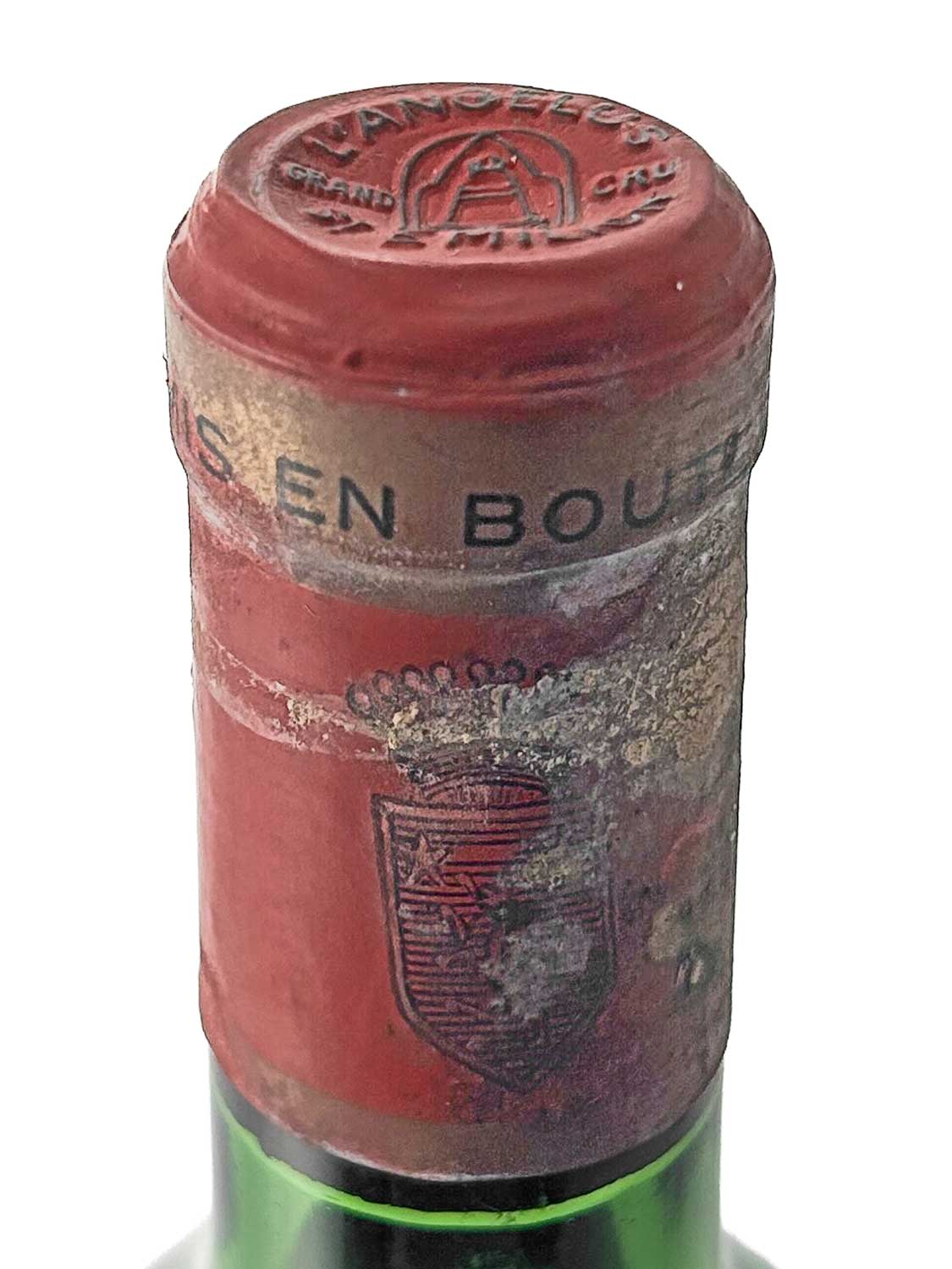 Nahaufnahme des Flaschenhalses einer Weinflasche mit rotem Kapselverschluss, auf dem oben 'ANGELUS GRAND CRU' und ein Glockensymbol geprägt sind, sowie ein teilweise beschädigtes Wappen auf der Vorderseite der Kapsel.