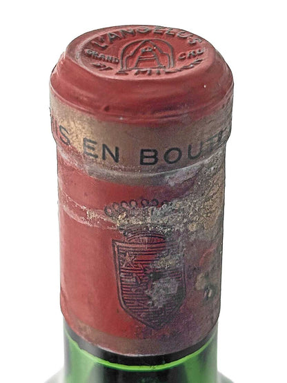 Nahaufnahme des Flaschenhalses einer Weinflasche mit rotem Kapselverschluss, auf dem oben 'ANGELUS GRAND CRU' und ein Glockensymbol geprägt sind, sowie ein teilweise beschädigtes Wappen auf der Vorderseite der Kapsel.