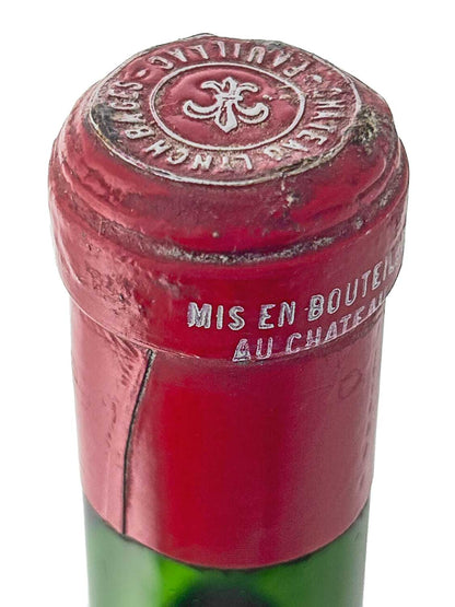 Flaschenhals mit rotem Kapselverschluss, oben geprägt mit dem Schriftzug 'CHÂTEAU LYNCH-BAGES' und einem stilisierten Kreuzsymbol, seitlich mit weißem Prägeaufdruck 'MIS EN BOUTEILLE AU CHÂTEAU'.