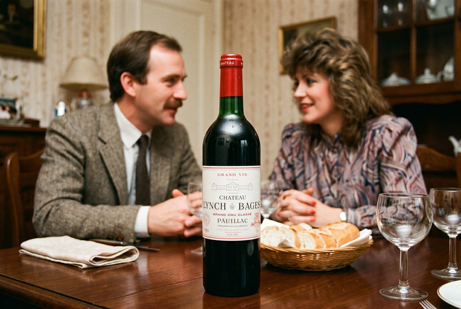 Flasche Chateau Lynch Bages 1985 auf einem Holztisch mit Brotkorb und zwei Personen im Hintergrund