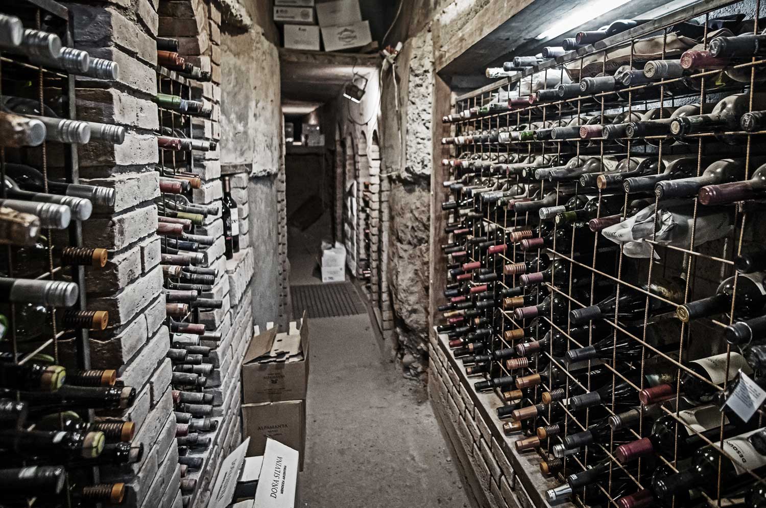 Weinkeller mit steinernen Wänden und Regalen, die zahlreiche Weinflaschen horizontal lagern, schmaler Gang mit Kartons auf dem Boden.